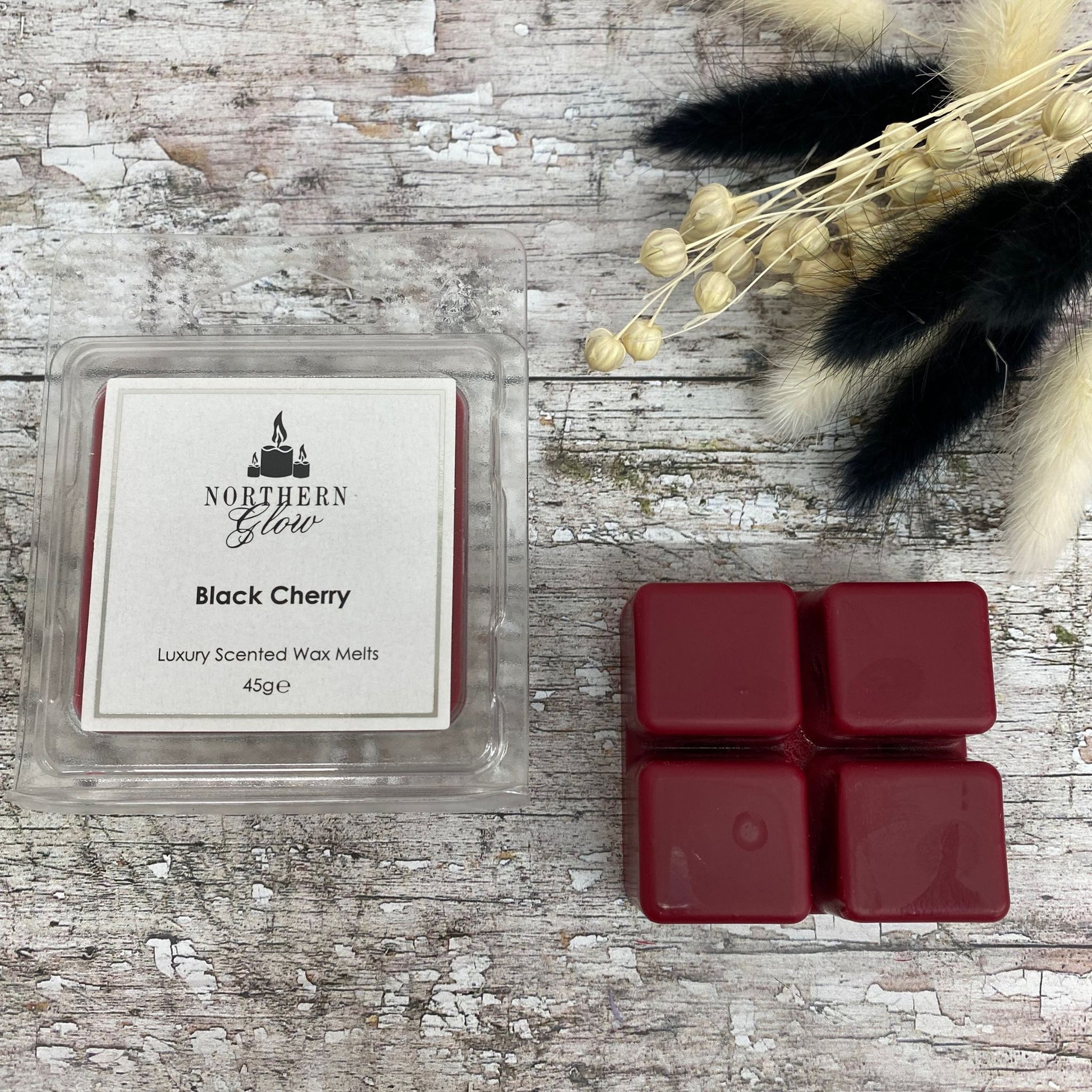 Black Cherry Wax Melt is a dark red colour shaped in a square with four breakable cubes in a plastic case. The label is white with a silver border and black writing. The logo is at the top with 'Black Cherry' in the centre.