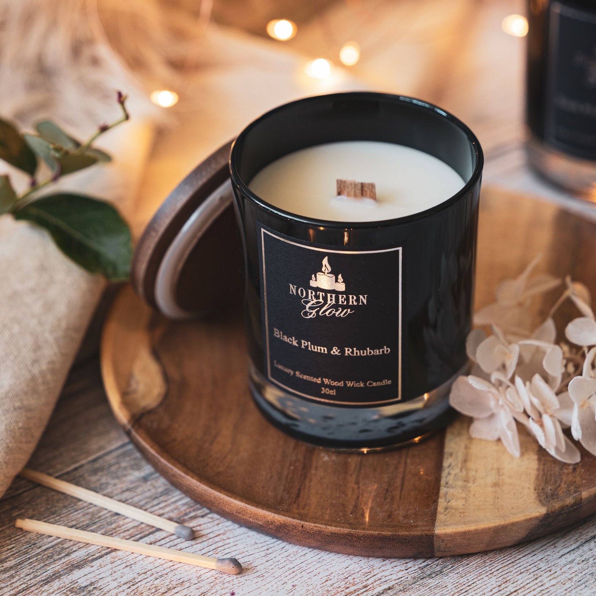 Black jar with a wooden lid standing against the jar. Wood wick is not lit, the label is black with silver writing and a silver logo and border. The scent is Black Plum and Rhubarb.