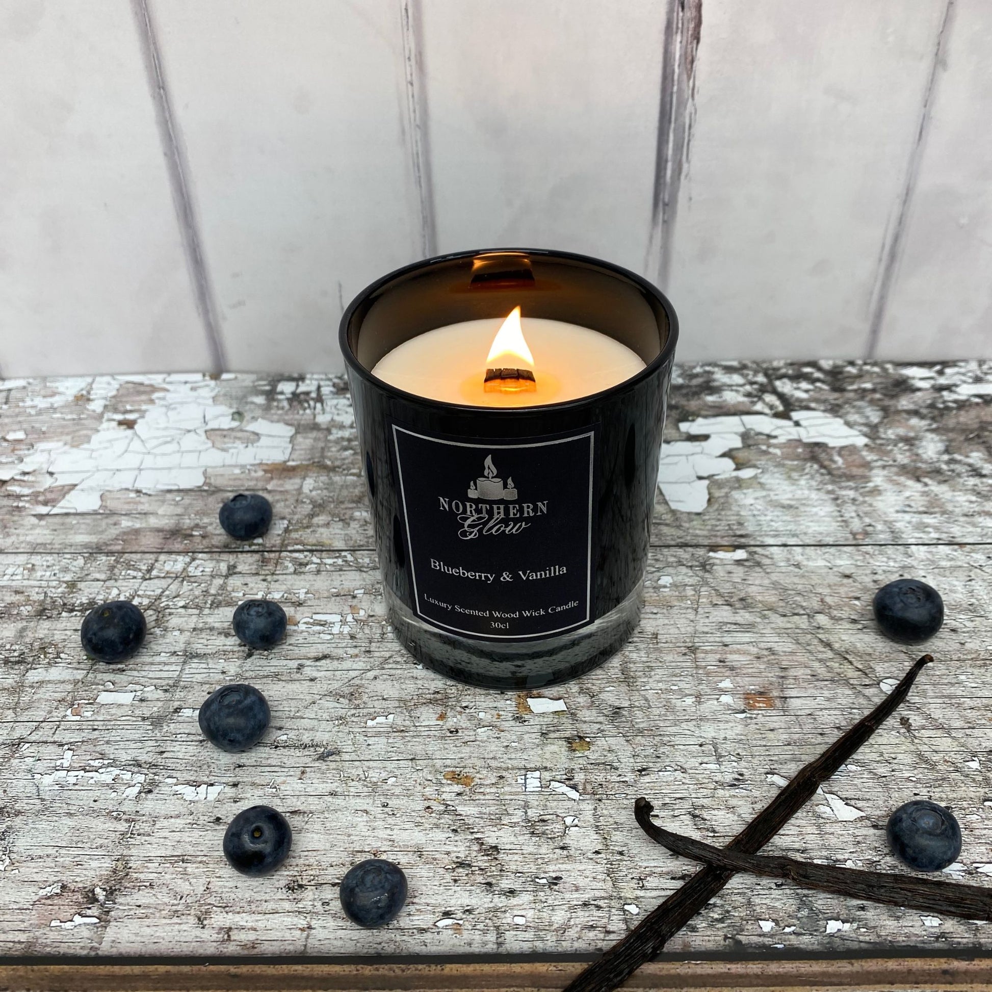 Blueberry and Vanilla scented wood wick candle is in a black jar with a black label and silver writing. The wood wick is lit with an orange flame. It has blueberrys and vanilla in the picture.