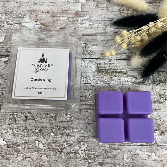 Cassis & Fig Wax Melt is a purple colour shaped in a square with four breakable cubes in a plastic case. The label is white with a silver border and black writing. The logo is at the top with 'Cassis & Fig' in the centre.