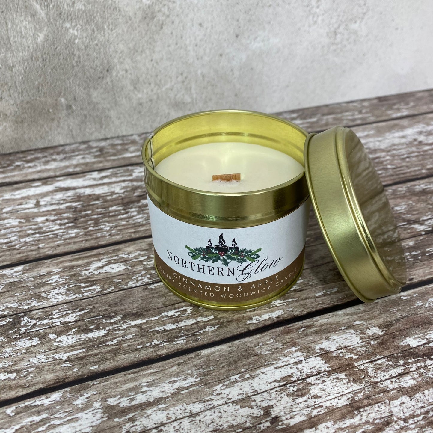 This wood wick candle is in a gold tin with a lid, the sticker is white with the logo in the centre. The writing is in white with a brown background with the words 'Cinnamon and Apple Scented Wood Wick Candle'