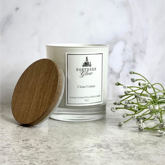 Clean Cotton Wood Wick candle is in a white jar with a white label with a silver border. It has a wooden lid and the logo is at the top with 'Clean Cotton' in the centre.