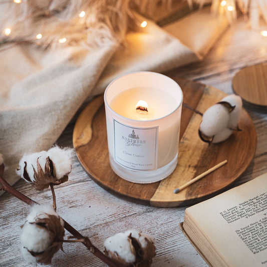 A white jar with the wood wick lit and a nice flame. It has a white label with a silver border, writing and logo. I t has balls of cotton in front of the jar and a wooden match.