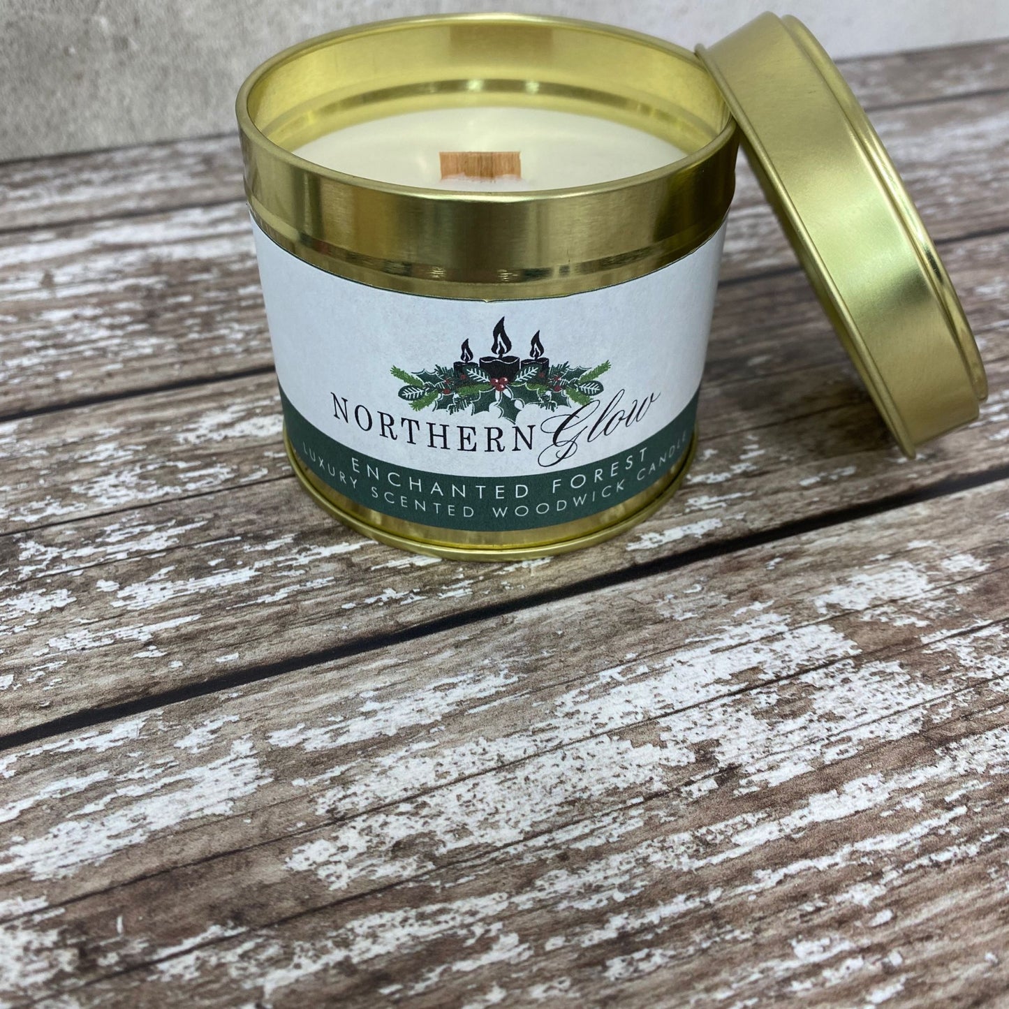 This wood wick candle is in a gold tin with a lid and it has a white label with the logo in the centre. The writing is white with a green background with the words ' Enchanted Forest Scented Wood Wick Candle'.