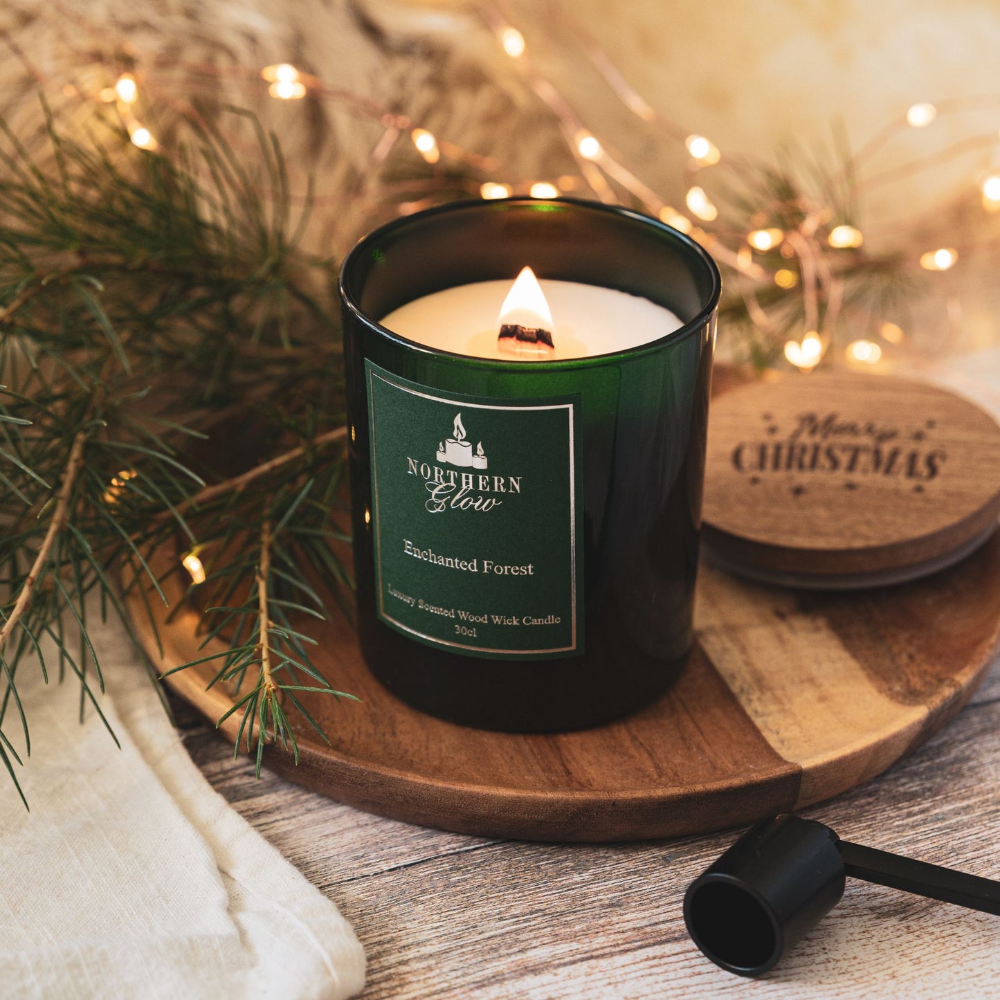 A green emerald jar with a wooden lid that has merry christmas written on the lid. The wood wick is lit and has a nice flame burning. It has a green label with a silver border, writing and logo.