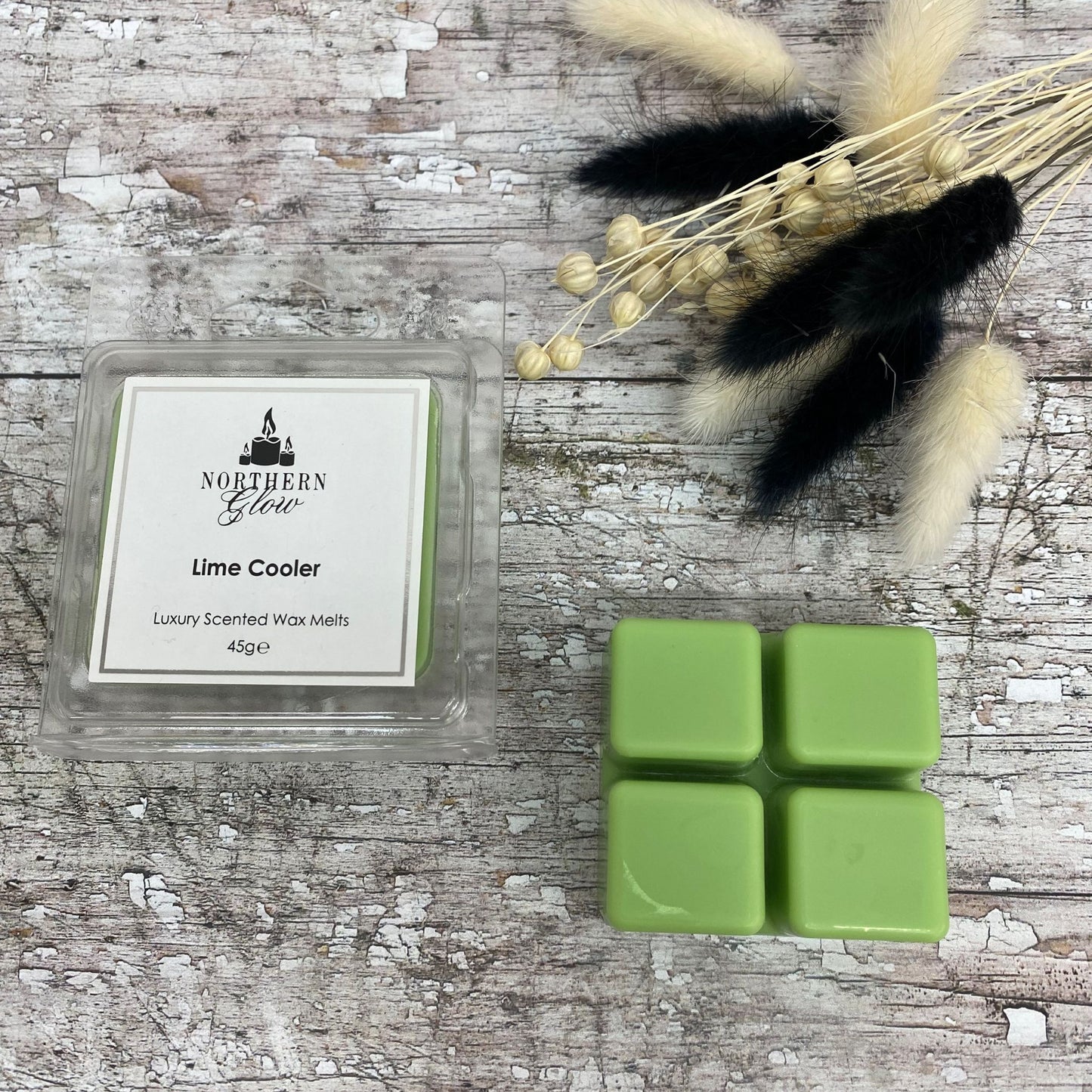 Lime Cooler wax melt is a green colour and shaped in a square with four breakable individual cubes in a plastic case. The label is white with a silver border and the logo is a t the top. The writing is in black writing with 'Lime Cooler' in the centre.