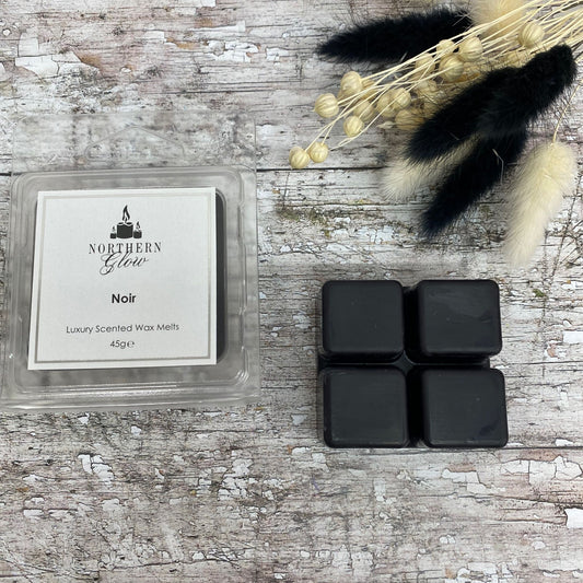 Noir Wax Melt is a black colour shaped in a square with four breakable cubes in a plastic case. The label is white with a silver border and black writing. The logo is at the top with 'Noir' in the centre.