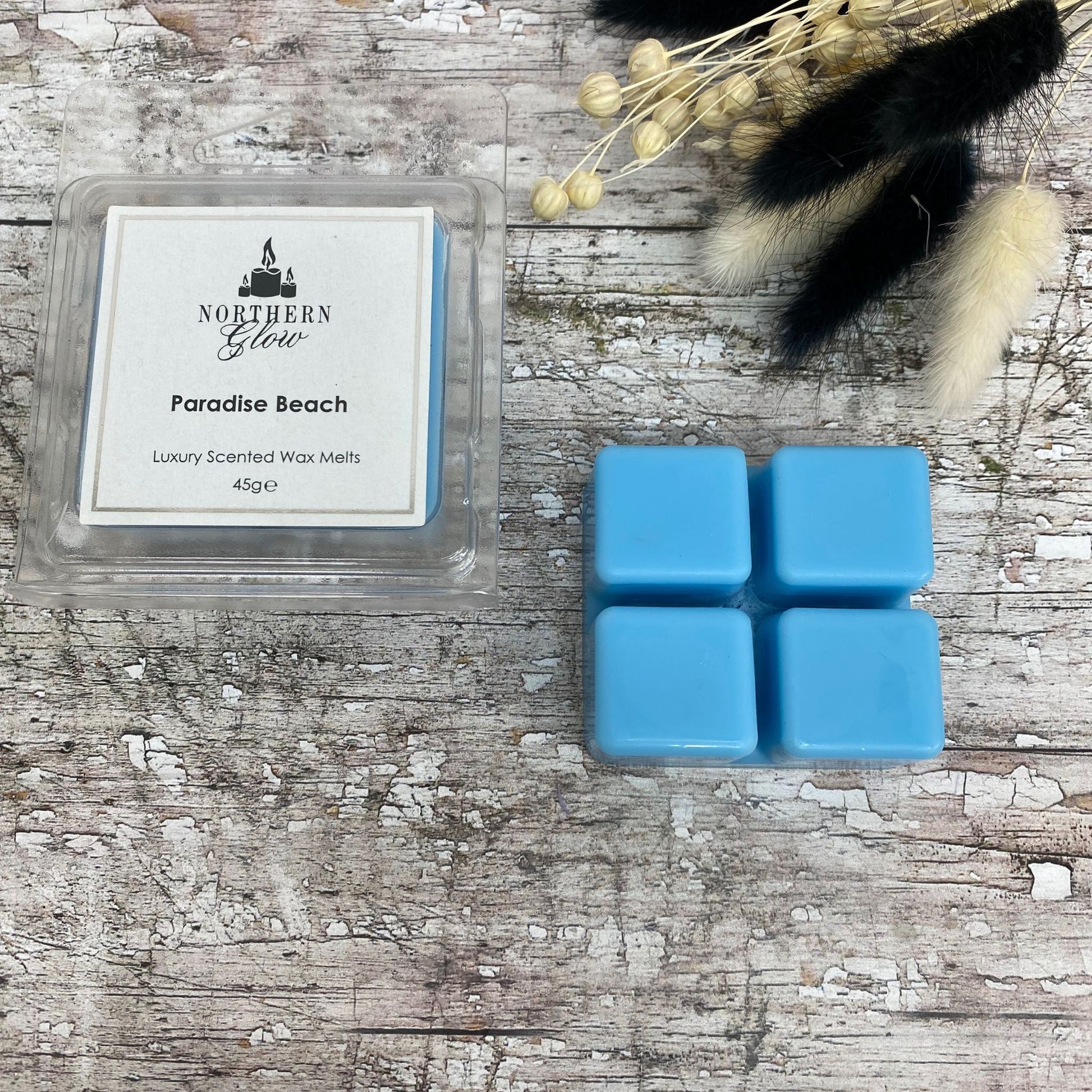 Paradise Beach Wax Melt is blue in colour and is square shaped with four breakable cubes in a plastic case. The label is white with a silver border and black writing. The logo is at the top with 'Paradise Beach' in the centre.