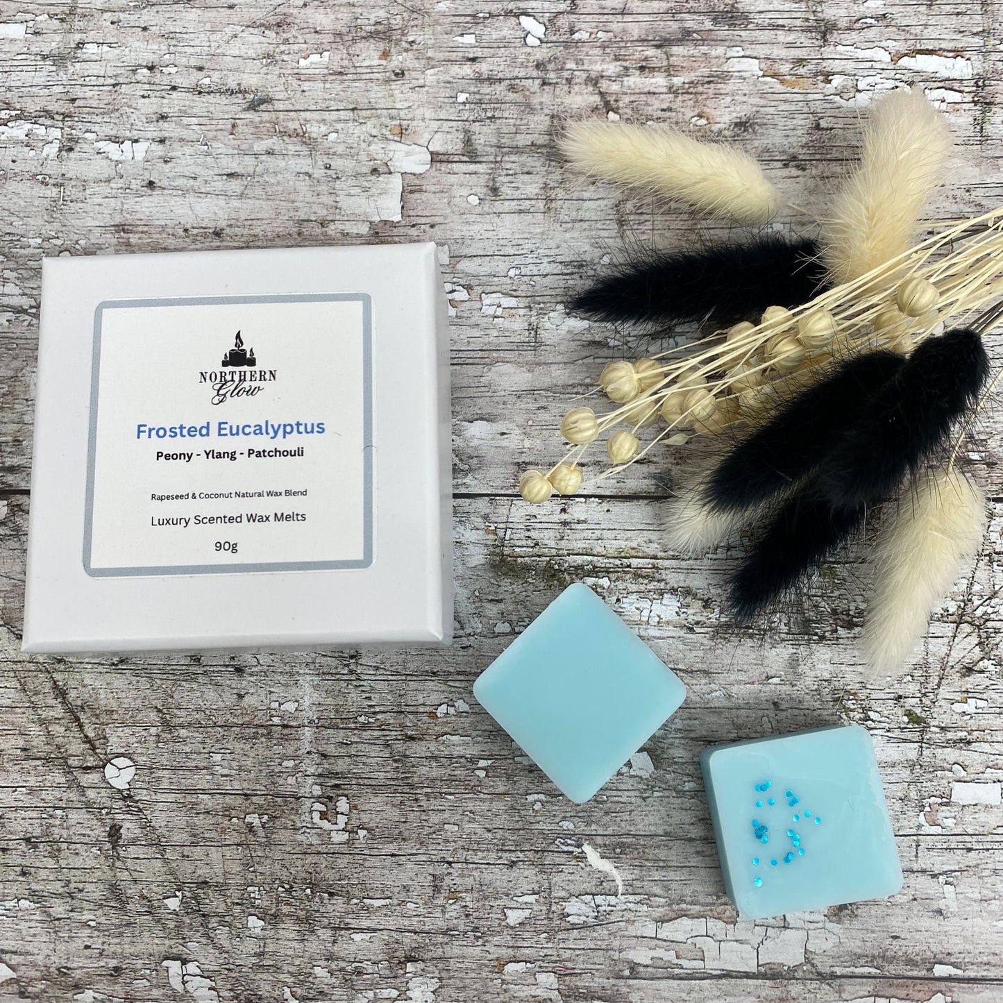 Frosted Eucalyptus wax melt is blue in colour and each melt is a square shape in a white box. There are eight individual wax melts in the white box. The label is white with a silver border. The logo is at the top with 'Frosted Eucalyptus' in blue writing in the centre.