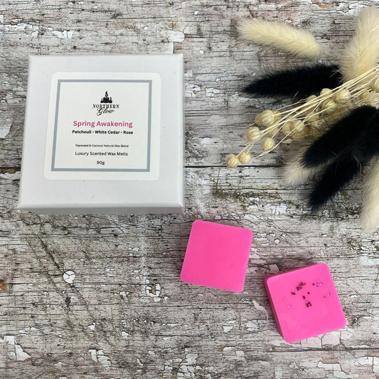 Spring Awakening wax melt is a magenta colour and each melt is a square shape in a white box. There are eight individual wax melts in the white box. The label is white with a silver border. The logo is at the top with 'Spring Awakening' in pink writing in the centre.