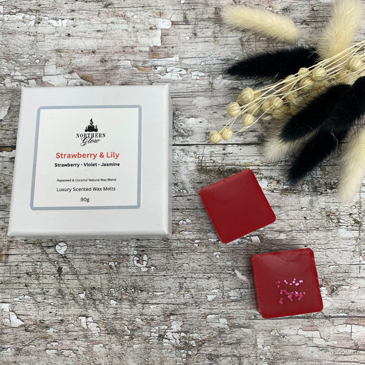 Strawberry and Lily wax melt is a red colour and each melt is a square shape in a white box. There are eight individual wax melts in the white box. The label is white with a silver border. The logo is at the top with 'Strawberry & Lily' in red writing in the centre