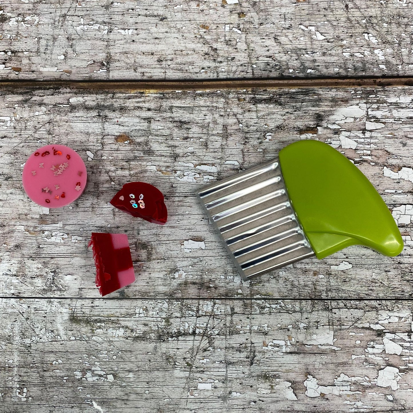 Wax Melt cutter is green with a silver cutter. This is used to cut chunks of wax melts. next to the cutter is a pink and red valentines cube that has been cut in two.