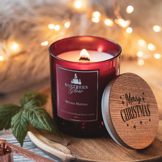 A red glass jar with the wood wick burning with a nice flame. It has a red label with silver writing, border and logo. It has a wooden lid against the jar that has Merry Christmas written on the lid.